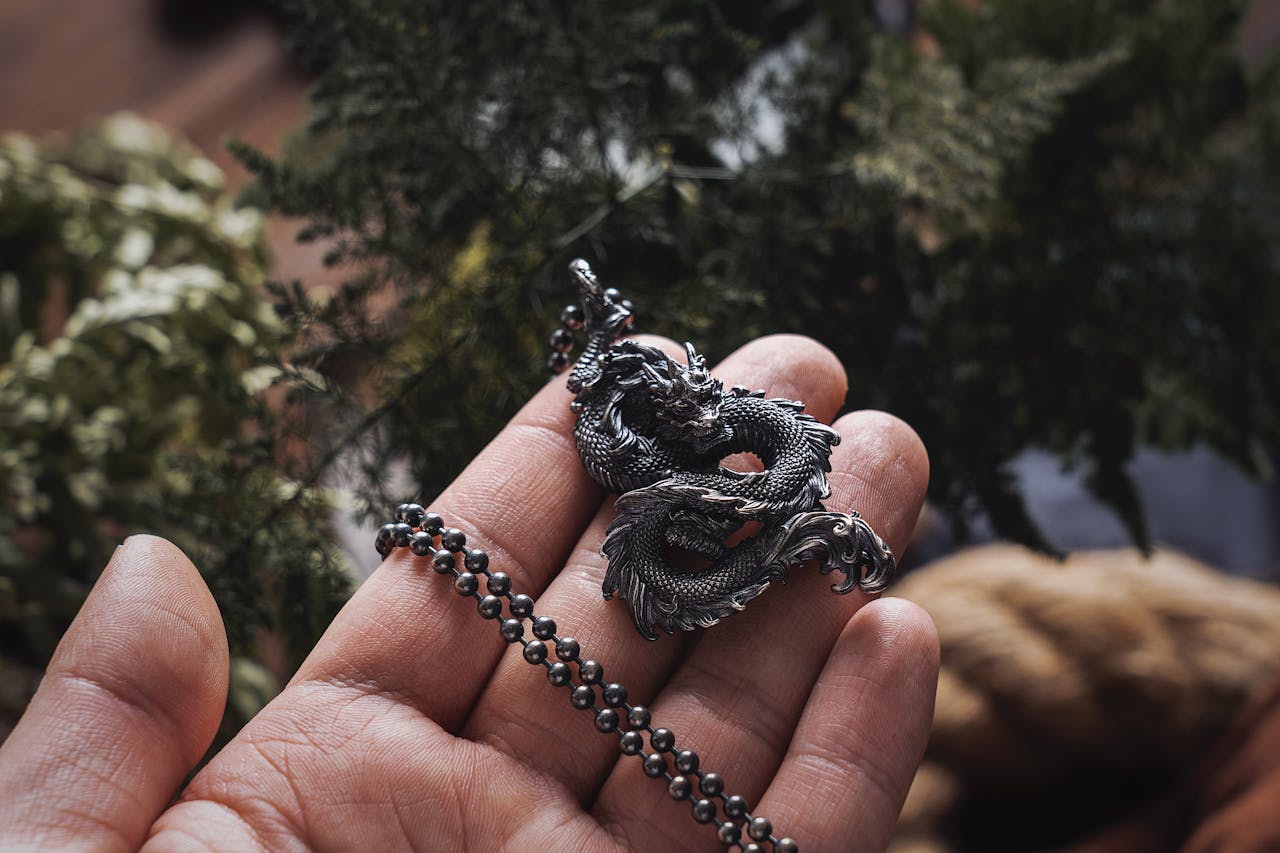 Close-up of a hand holding an ornate Chinese dragon necklace, surrounded by greenery.